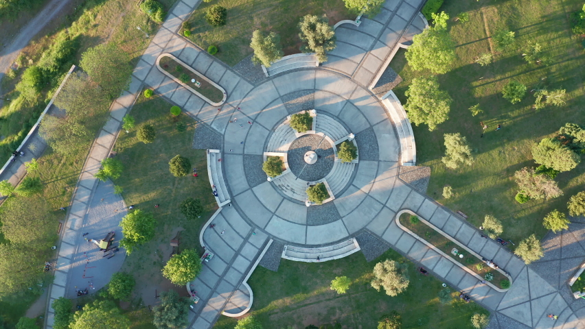 Round paved playground in a garden surrounded by trees and grass, as the central square of green city park and recreational area in urban environment. Aerial drone view of walkways and podium.