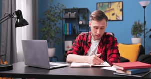 A focused college student boy is video chatting on a laptop. A male teenager in a flannel red shirt works remotely while sitting at a desk in the living room. Young man gestures while video chatting.