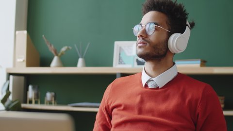 Tilt up shot of young handsome mixed race businessman putting on wireless headphones and listening to music while sitting at workplace in office