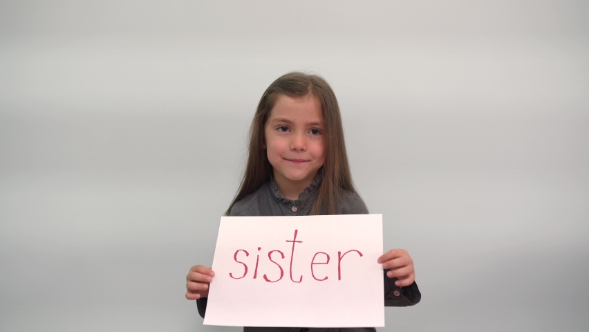 A girl holds a piece of paper with the inscription SISTER and smiles A modest child shows a sign with the word SISTER on a gray-blue background Cheerful children Happy family