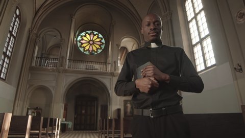 Low angle view of young bald African-American priest wearing black clothes and collar standing in Christian church with Bible in hands and looking at camera