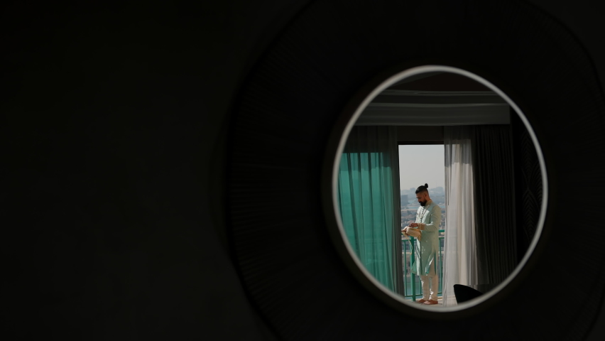 Young Arab In Blue Suit With Embroidery. Man With Beard And Stylish Hairstyle. Man Wears An Arabic Headdress. View Through Small Round Window. Man Standing On The Balcony.