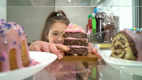 Little girl opening fridge door and taking out a piece of cake and a lot of sweets. Point of view from inside of the kitchen refrigerator