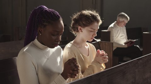 Medium shot of two diverse young women sitting on wooden bench in Lutheran church with hands folded for prayer and closed eyes and praying during Sunday service