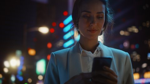 Beautiful Woman Standing, Using Smartphone on a City Street with Neon Bokeh Lights Shining at Night. Smiling Beautiful Female using Mobile Phone. Medium to Close-up Dolly Cinematic Flare Shot: stockvideo