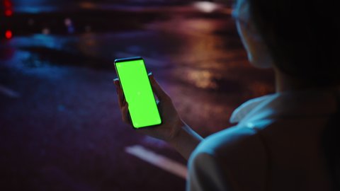 Beautiful Woman using Chroma Key Smartphone while Walking Through Night City Street Full of Neon Light. Female Using Green Screen Mobile Phone. Over the Shoulder Tracking Medium Shot