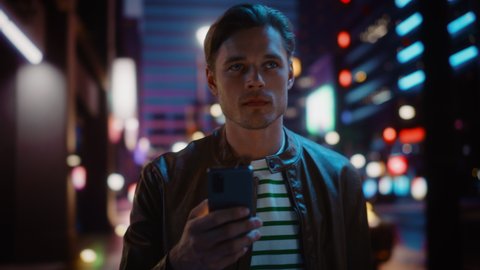 Man Using Smartphone Walking Through Night City Street Full of Neon Light. Smiling Stylish Man Using Mobile Phone, Social Media, Online Shopping, Texting on Dating App. Dolly Tracking Medium Shot