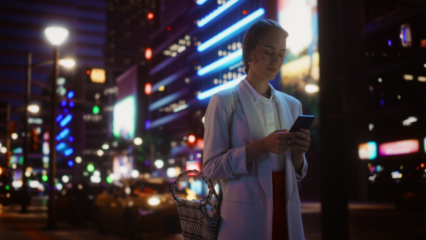 Beautiful Young Woman Using Smartphone Walking Through Night City Street Full of Neon Light. Smiling Thoughtfully Female Using Mobile Phone, Posting Social Media, Online Shopping, Texting. Low angle Royalty-Free Stock Footage #1072234943