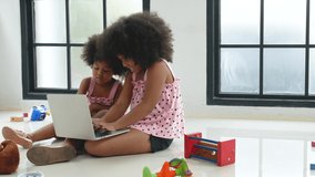 Two African American little girl enjoy to play notebook and sit on the floor among several types of toys in living room and they look happiness.