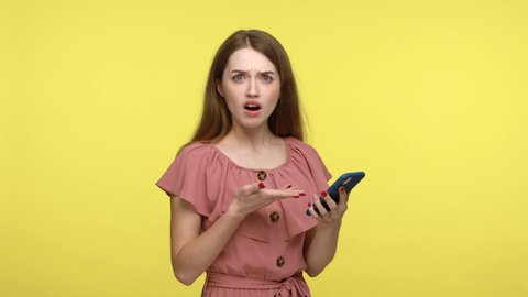 Extremely surprised brown haired woman in pink dress looking smartphone display with astonishment, receiving message with shocking breaking news, Indoor studio shot isolated on yellow background.