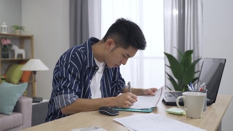 tired asian male doing paperwork is putting down the pen to massage aching lower back and exercising stiff neck with a painful look in the living room at home.