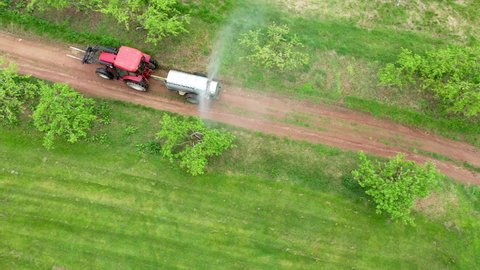 Aerial looking down onto tractor pulling fan sprayer spraying bugs in orchard.