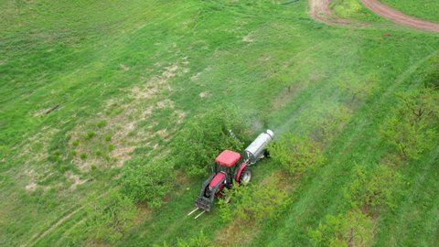 Aerial in front of tractor spraying fruit tress with fan sprayer in orchard.