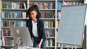 Distance training, online lecture. Young smart african american female teacher conducts online lesson for students via video conference uses computer, stand at library, shows information on whiteboard - Powered by Shutterstock - Get 15% off with code: PIKWIZARD15