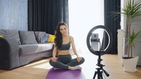 Beautiful woman in sportswear sitting in lotus posture and recording video with smartphone on tripod. Social media influencer waving goodbye to followers and switching off camera. Concept of blogging