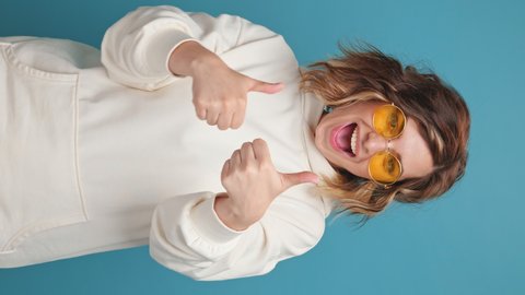 Vertical video stories reels attractive young woman face close up showing Like sign smiling with hands gesture body language looking camera wearing sunglasses curly hair on blue background Positive