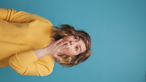 Vertical video stories reels frustrated pretty young woman yawns covering her mouth with her hand with displeased face from boredom curly hair yellow clothes on blue background. Emotion Lifestyle