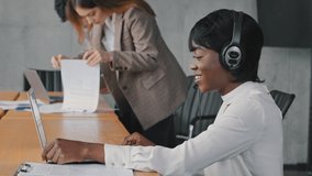 Young african business woman working call center manager support line agent speaks remote communication wears headset headphones advises client makes videocall in office on background of colleagues