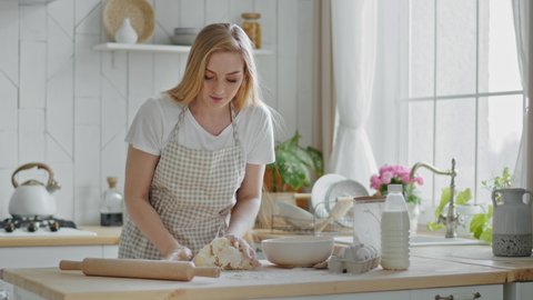 Adult beautiful woman 35s housewife baker mother wears apron cooking stands in kitchen preparing bread homemade pastries biscuits kneads dough with wheat flour on wooden table heaving rolling pin