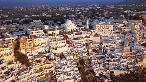 Aerial Panning Shot Of Orthodox Metropolitan Cathedral In City, Drone Flying Backward From Structures On Mountain At Sunset - Santorini, Greece