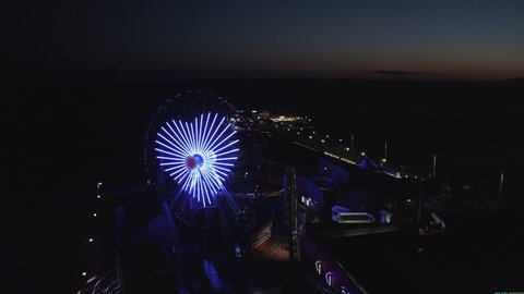 Aerial Lockdown Shot Of Illuminated Heart Shape On Ferris Wheel At Pier, Drone Flying Over Famous Santa Monica Pier At Night