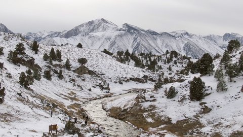 Aerial Shot Of Creek Amidst Snowcapped Mountain Range, Drone Descending Forward Towards Tourists Hiking At Hot Creek Geological Site - Mammoth Lakes, California