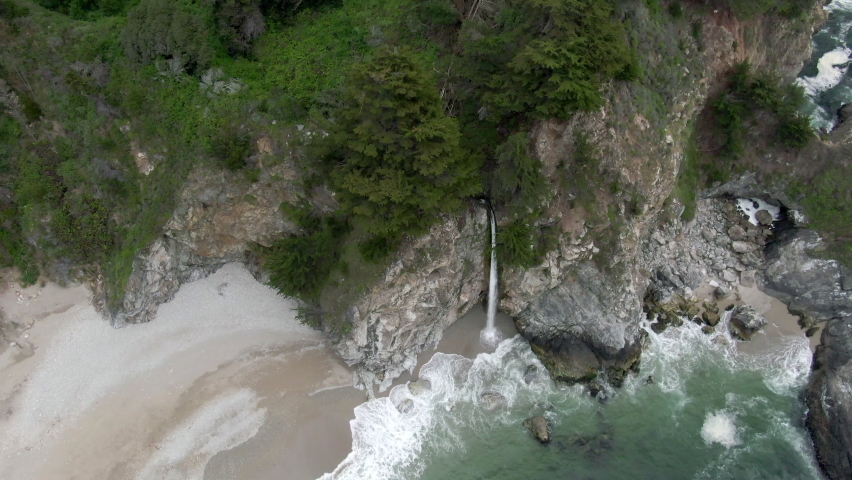 Aerial Tilt Up Shot Of Mcway Falls From Rocky Cliff At Beach, Drone Flying Backward Over Waves Splashing On Shore - Big Sur, California