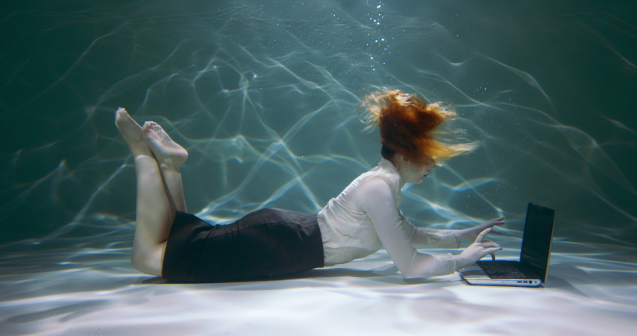 Cinematic shot, young redhead business woman using laptop under water to consult customers, swimming up slow motion.