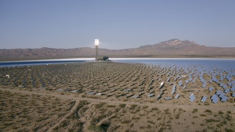 Aerial Reversing Away From A Solar Field In The Desert With Dirt, Dry Brush, Clear Blue Sky, Bright Sunlight, And Steep Mountains In The Background - Ivanpah, California