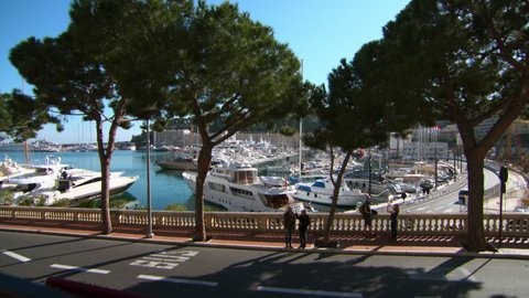 Lockdown Of A Bridge And Overpass Overlooking Monte Carlo Harbor With Moored Boats, Trees, And Cars Driving Along Coastline Street