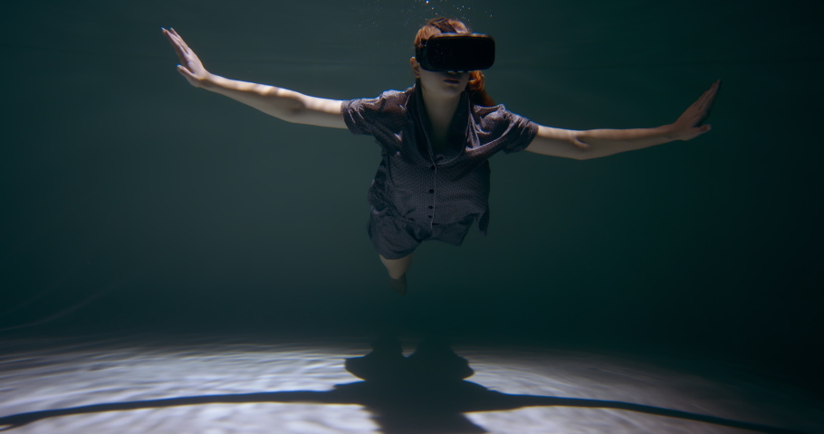Young beautiful redhead woman wearing night pyjamas using VR headset deep under water on dark pool floor slow motion.