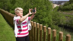 Senior woman doing panoramic video with cellphone at park on sunny day. Elder lady taking photo with smart phone on natural landscape