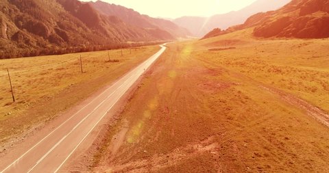 Aerial UHD 4K view. Low air flight over mountain asphalt highway road and meadow at sunny summer morning. Near green trees. Fast horizontal movement with sun rays.