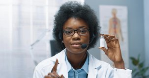 Close up of young joyful African American female physician sitting at desk in hospital cabinet talking in headset and showing covid-19 vaccine, coronavirus cure, online video consultation
