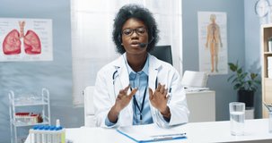 Portrait of cheerful young pretty African American female doctor specialist sitting at table in clinic and speaking in headset, medical webinar teaching medicine online video consultation with patient