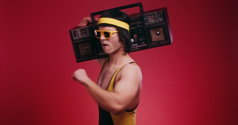 Funny retro athlete man in yellow tracksuit and sunglasses from 80's with ghettoblaster dancing and turning around on red background. Retro sport hipster show off and posing in front of camera.