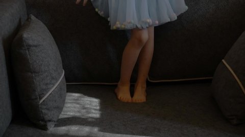 Toddler girl in blue tulle tutu skirt with barefoot legs jump on couch with pillows. Girl kid having fun at home and playing and dancing like princess
