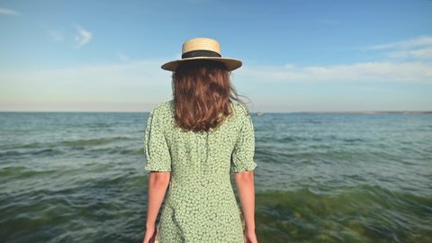 Close-up back view young attractive slender Caucasian woman in a green dress and a straw hat walks in the summer along a wooden pier on the seashore. Relaxation and relaxation on vacation and travel