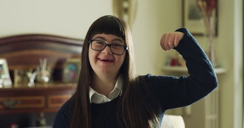 Authentic shot of happy friendly teen girl with down syndrome showing her power with arms and smiling in camera at home. Concept of persons with disabilities, child care, healthcare, people, optimism