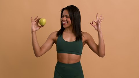 Healthy Weight-Loss Diet. African Fitness Woman Posing With Apple Gesturing Okay Smiling To Camera Approving Weight Loss Nutrition Standing On Beige Studio Background. Dieting And Slimming Concept