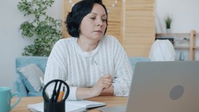 Mature woman is talking using laptop to make online video call and taking notes indoors at home. Distant work and internet communication concept.