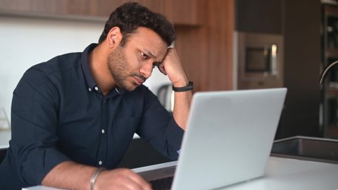 Upset multi-ethnic man is using laptop sitting at home alone, pensive young indian guy sits in the kitchen at with the laptop, rested head with hand, has financial problems and issues with project
