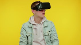 Teen stylish boy using virtual reality futuristic technology VR app headset helmet to play simulation 3D 360 video game, drawing. Young man on yellow studio background. People sincere emotions