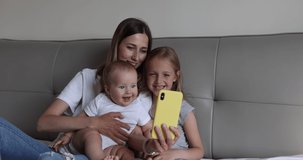 Happy caucasian mother and kids have online video call with father or grandmother via mobile phone when lying on bed in cozy bedroom at home. Slow motion