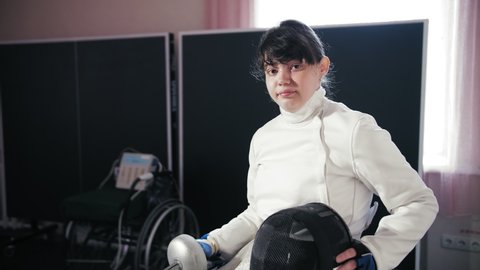 disabled athlete. fencing. paralympic games. wheelchair fencing. Portrait of disabled female fencer, swordsman sitting in wheelchair, wearing white fencing costume, uniform and holding black fencing