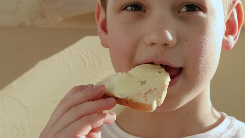 The boy with shutter yeses in white shirt holding a toast with coconut paste with pleasure. An allergic child with red cheeks eats white bread. Royalty-Free Stock Footage #1072317317