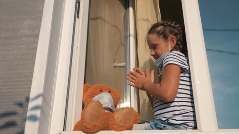 girl in medical mask sits by an open window and applauds. child thanks medical staff fighting Covid-19 infection. little girl applauds her hands to doctors on street, looks out window wearing a mask.