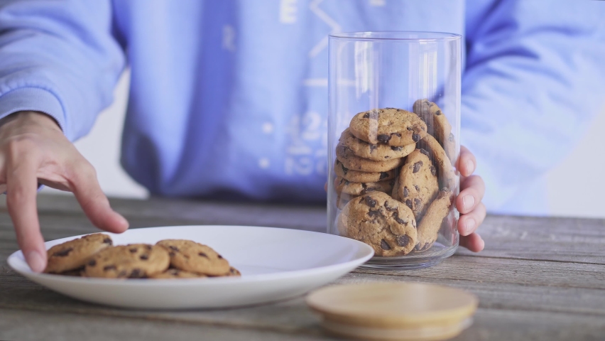 Homemade baking. The girl puts cookies from a white plate into a glass jar. Chocolate chip cookies in a glass jar with a bamboo lid. Close-up.