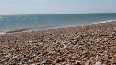 Pebble British beach, selective focus. Pebble colorful coastline, horizontal. Waves are reaching pebble beach. Sky, horizontal line, waves and pebbles. Dungeness Headland, Kent, England.