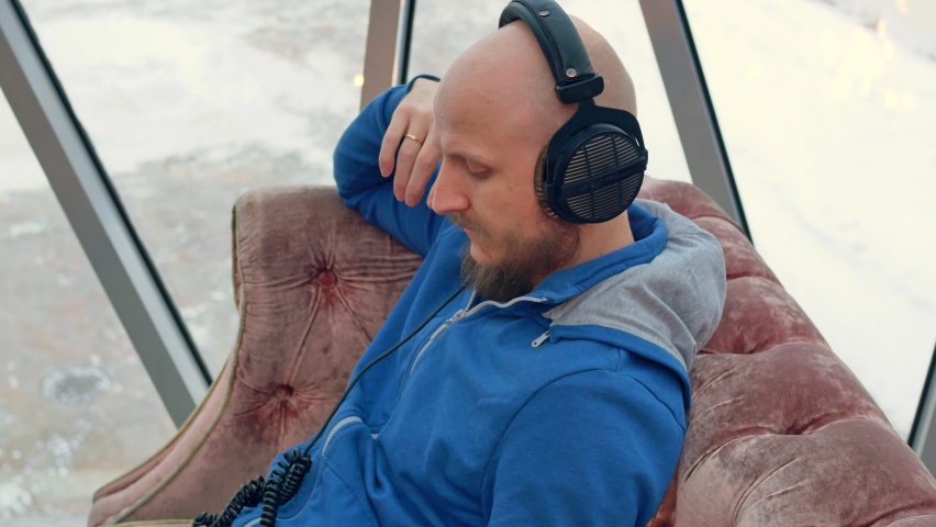 A bald young man with an attractive beard and a beautifully styled mustache listens to music with professional headphones on a velor sofa near the window.